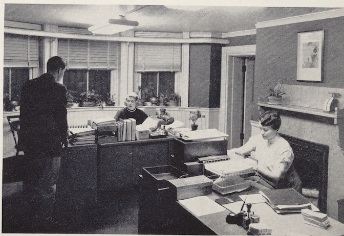 A view of an office with two desk and people working.